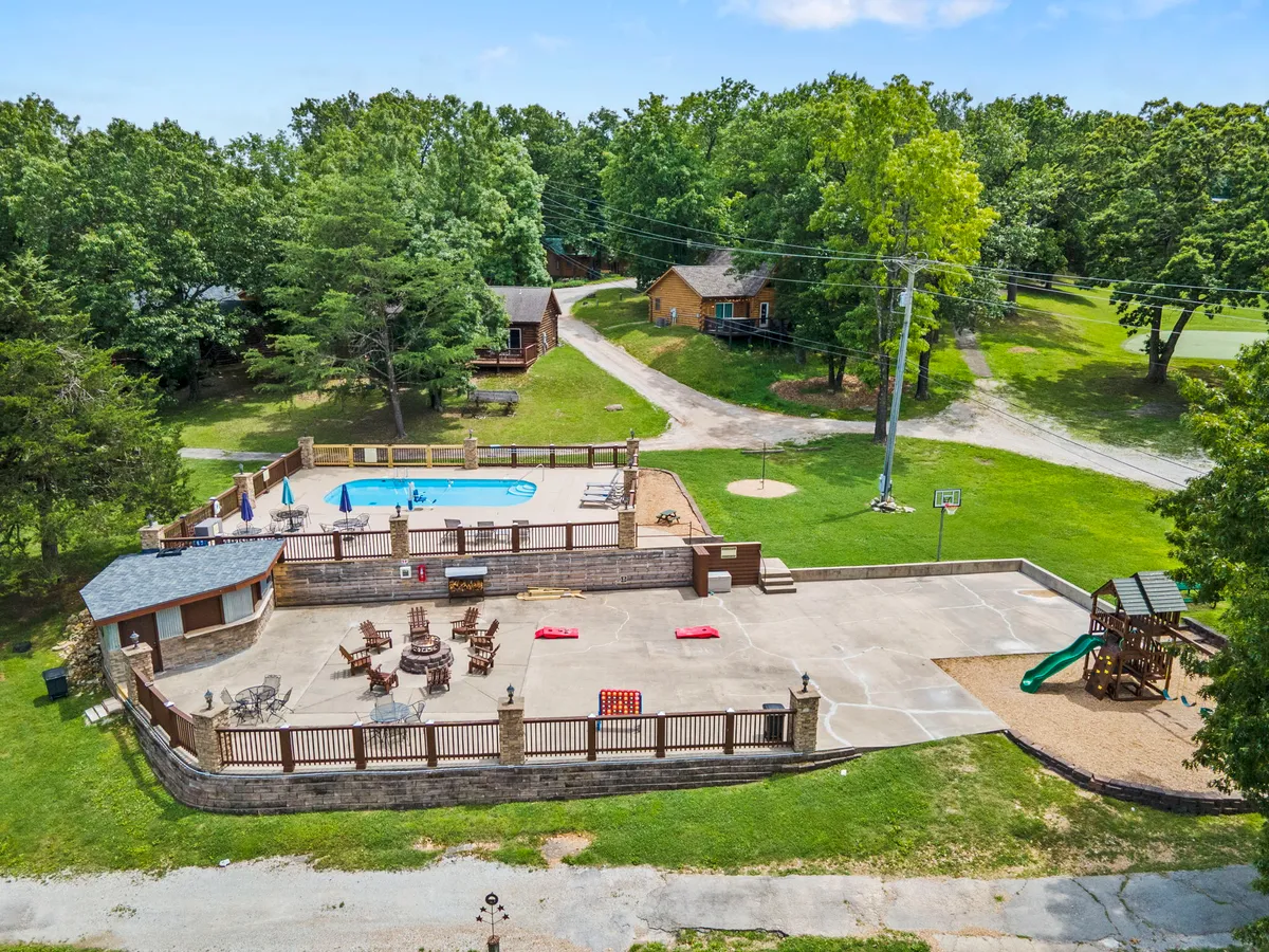 This image captures a vibrant resort hub, featuring a swimming pool, patio area, playground, seating, and a basketball hoop, all set against a backdrop of lush greenery. Children are visible on the playground, while families relax by the pool and others engage in outdoor games.