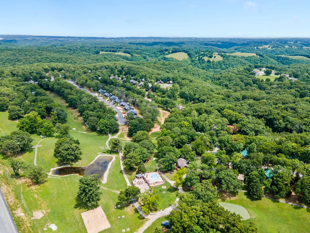 A wide view of the resort grounds featuring an inviting swimming pool, expansive green lawns, scenic hills, a pond, and a cluster of cabins. Guests can be seen enjoying various outdoor activities like swimming and nature walks.