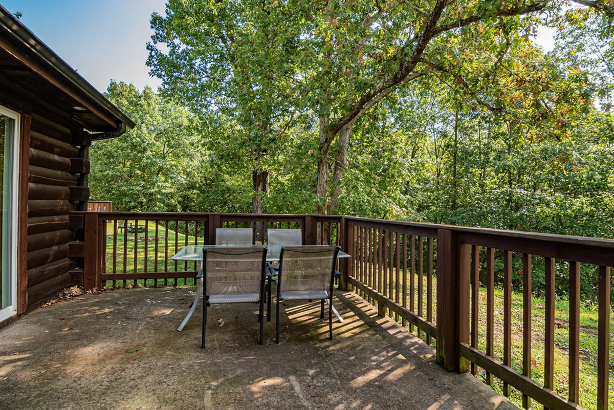 A spacious wooden deck attached to a log cabin, outfitted with a dining table and chairs, bordered by lush greenery. The area is perfect for al fresco meals and soaking up the tranquil outdoor atmosphere.