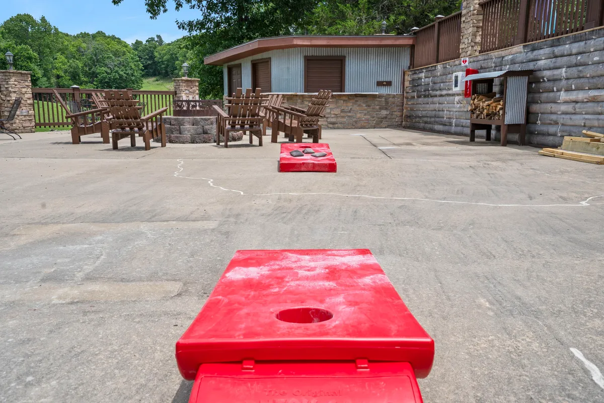 An inviting fire pit area, complete with red cornhole boards, wooden chairs, and a stone wall, awaits guests for casual evening gatherings. The setup suggests friendly games, cozy fireside chats, and relaxation in the fresh air.
