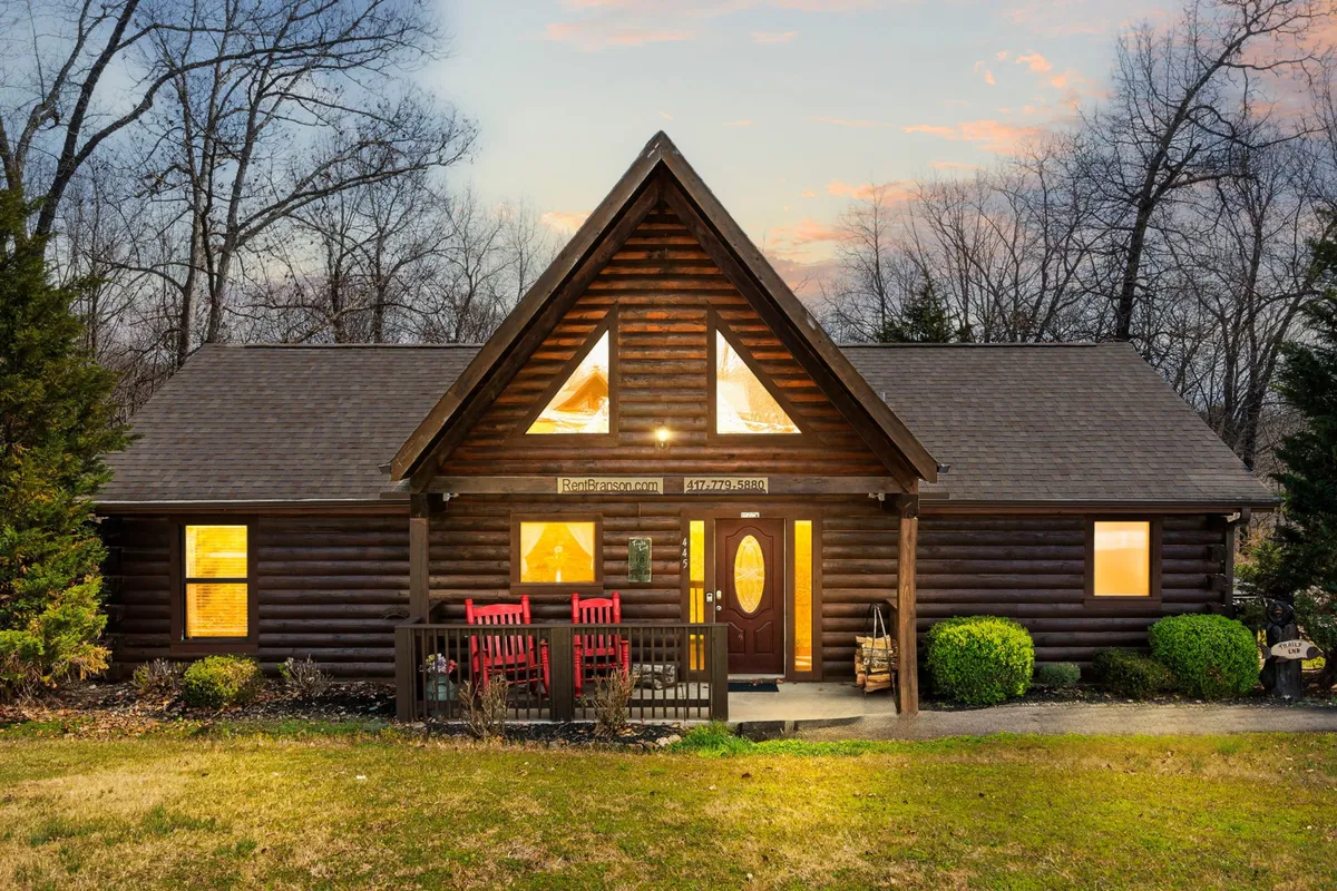 A classic log cabin nestled among trees, with inviting red rocking chairs perched on a spacious front porch and large triangular windows reflecting the forest surroundings. The porch offers a perfect spot to sip coffee while immersed in the quiet seclusion of the woods.
