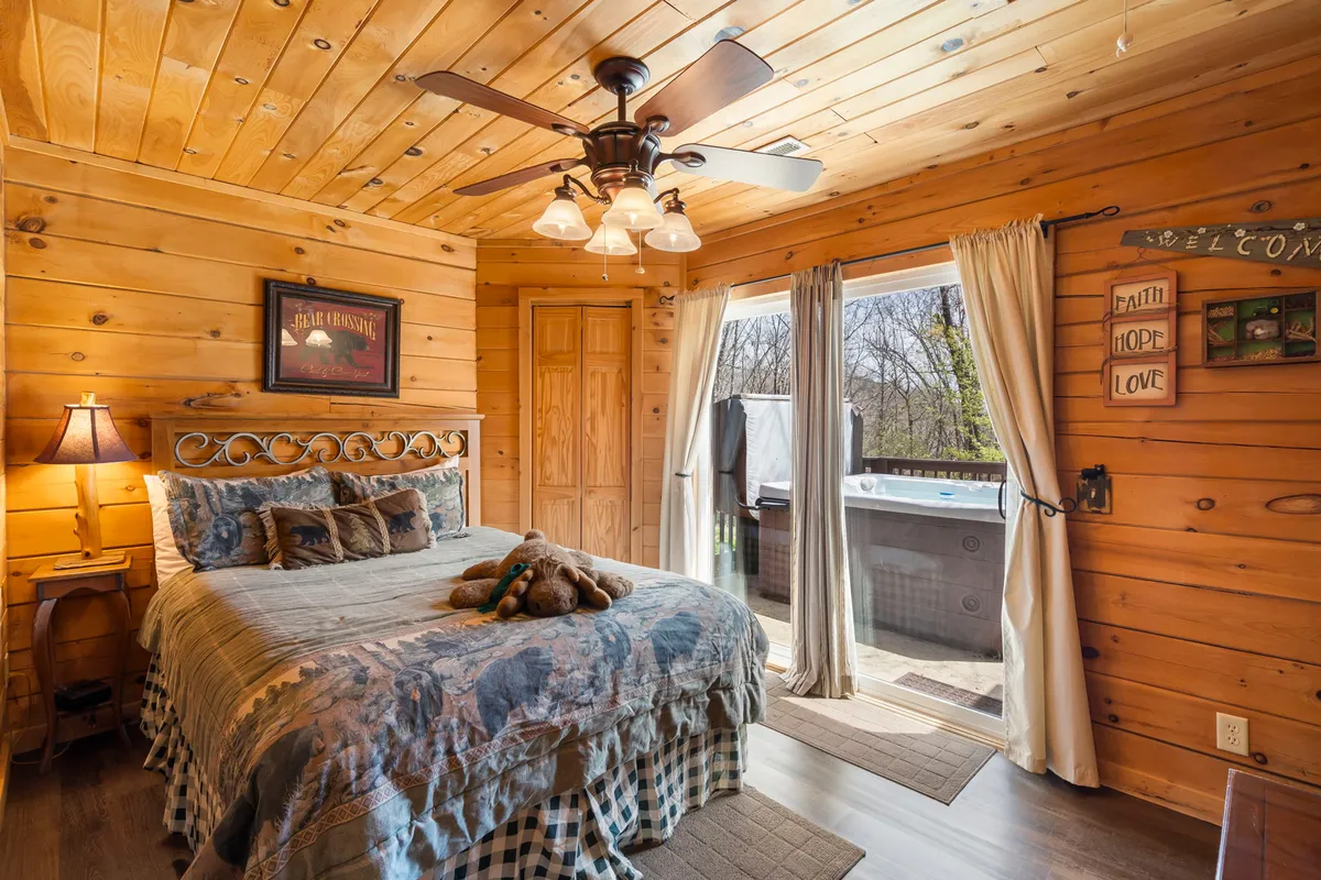 A warmly lit bedroom with a plush king bed, wooden ceiling, and an open door leading to an outdoor hot tub. The scene exudes comfort and privacy, with hints of nature visible through the doorway, suggesting a seamless transition between indoor luxury and outdoor relaxation.