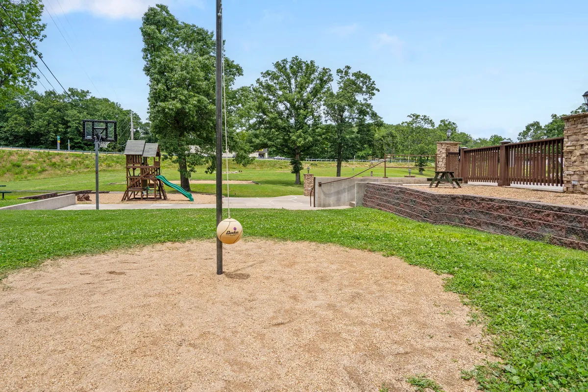 An exterior recreation area featuring a volleyball pole, basketball hoop, and playground structure. Children are playing on the equipment while families gather nearby, suggesting an atmosphere of fun and friendly competition.