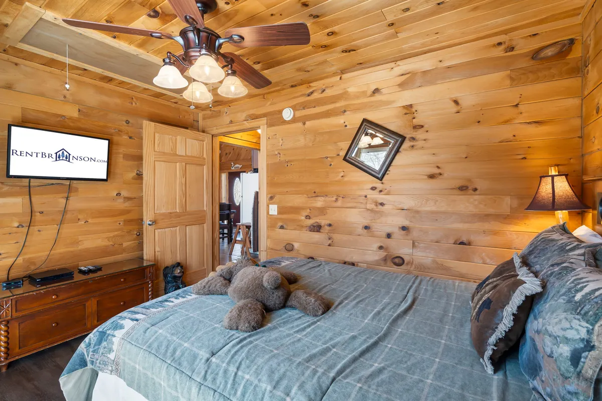 A cozy bedroom with wooden walls, a bed adorned with plush decorative pillows, a teddy bear, and a TV mounted on the wall. Warm lighting and soft textiles create an inviting, homey vibe perfect for unwinding.