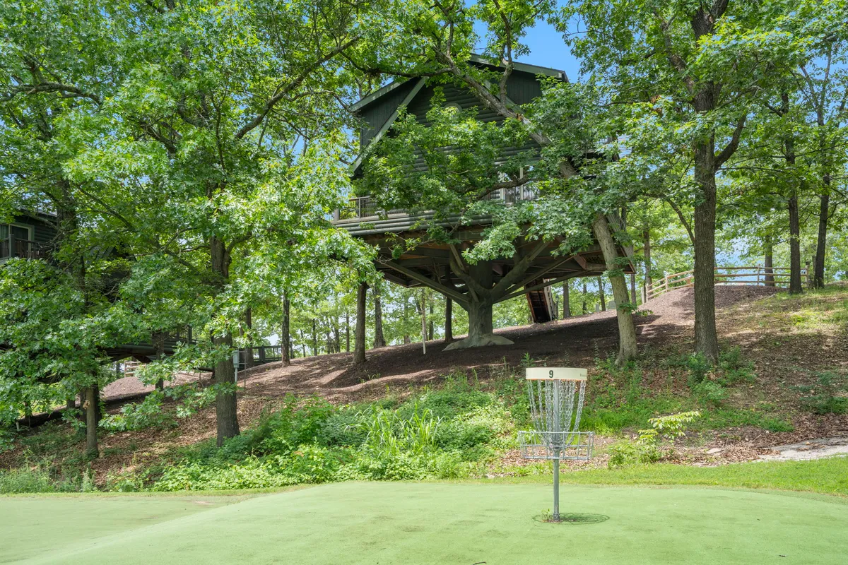 The image features a disc golf basket situated near a playful treehouse, surrounded by lush greenery. The area beckons for outdoor adventure and friendly competition, capturing the spirit of the 27-hole disc golf course within the resort.