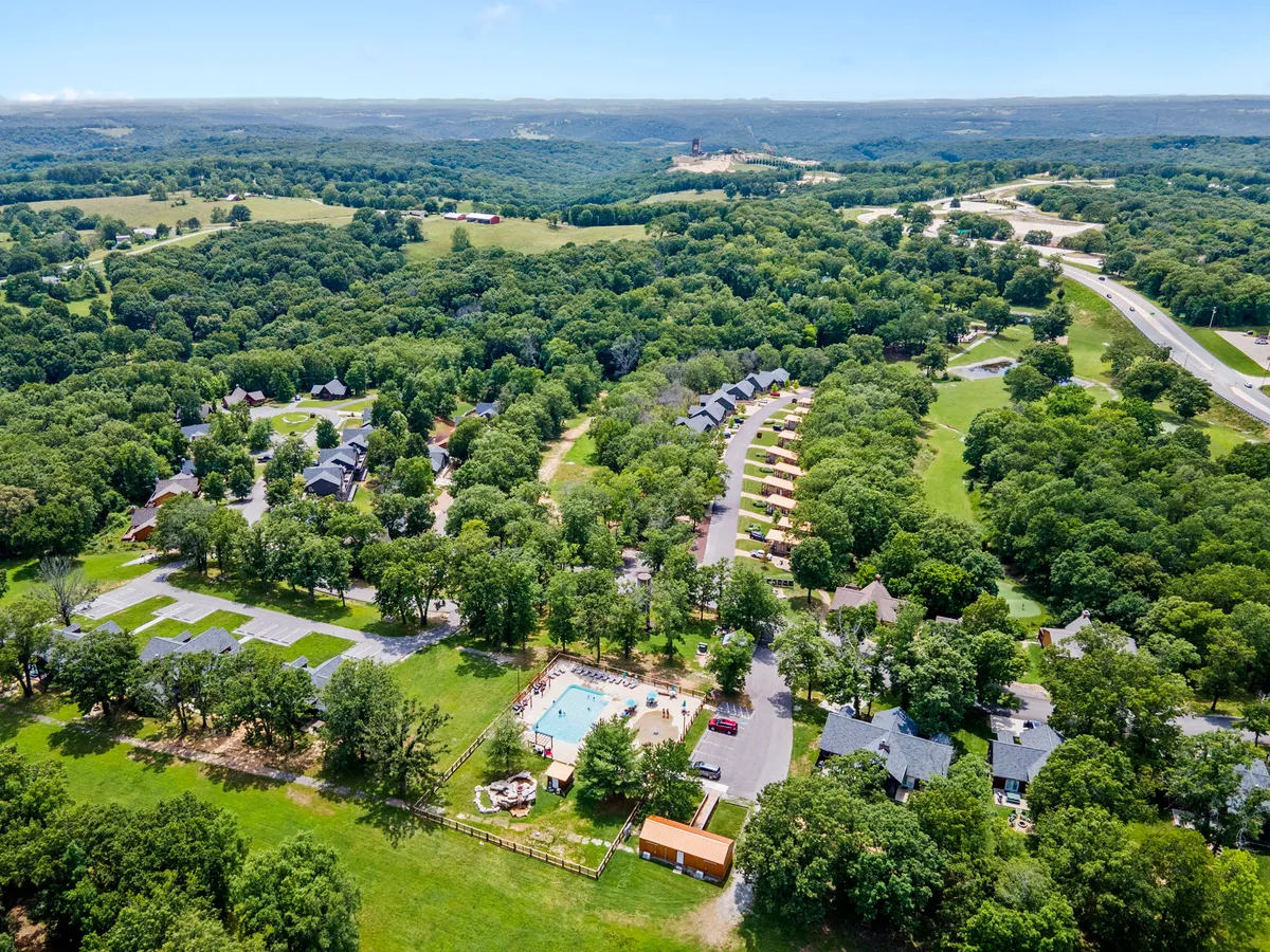 A wide exterior shot shows a swimming pool surrounded by cabins nestled in a lush, green landscape. The pool area is lively, suggesting a hub for swimming, picnicking, hiking, and family gatherings. Trees and open space reinforce the feel of a nature-rich, activity-filled resort environment.
