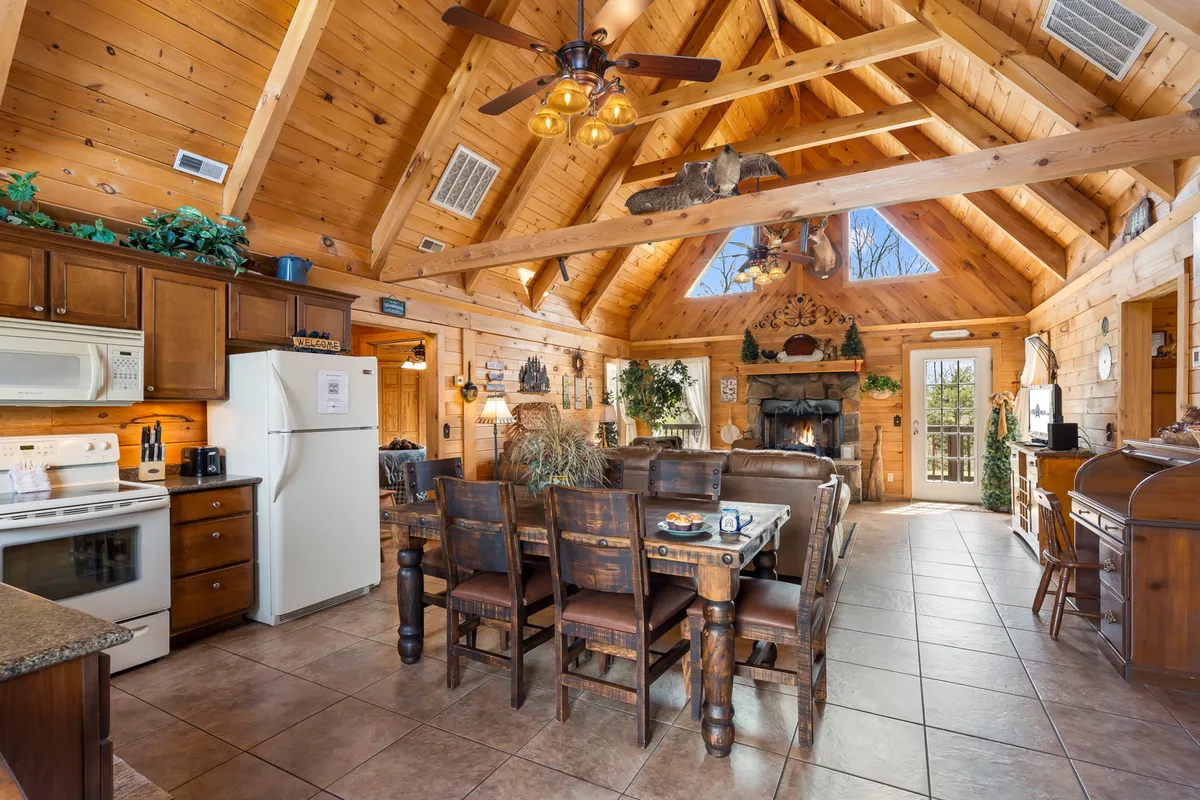 A spacious, open-concept area displaying the dining table, wooden beams, a fireplace, kitchen appliances, and generous windows. The scene captures family and friends sharing meals, the convenience of a well-equipped kitchen, and the inviting ambiance of the dining and living area combined.