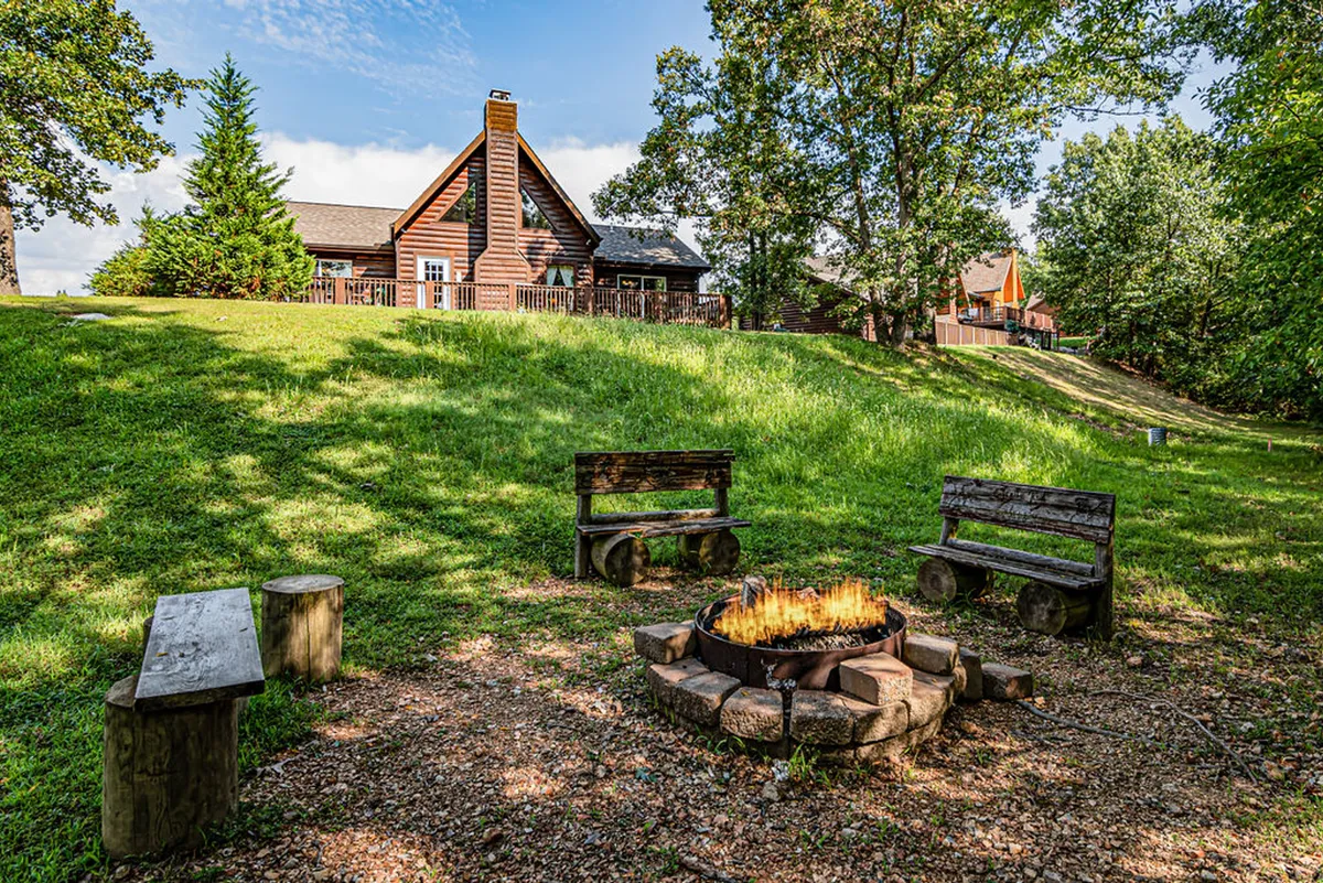 A cozy fire pit area with wooden benches set in green grass outside a rustic log cabin, framed by trees. The setup invites guests to gather for stories, s'mores, and stargazing under the open sky.