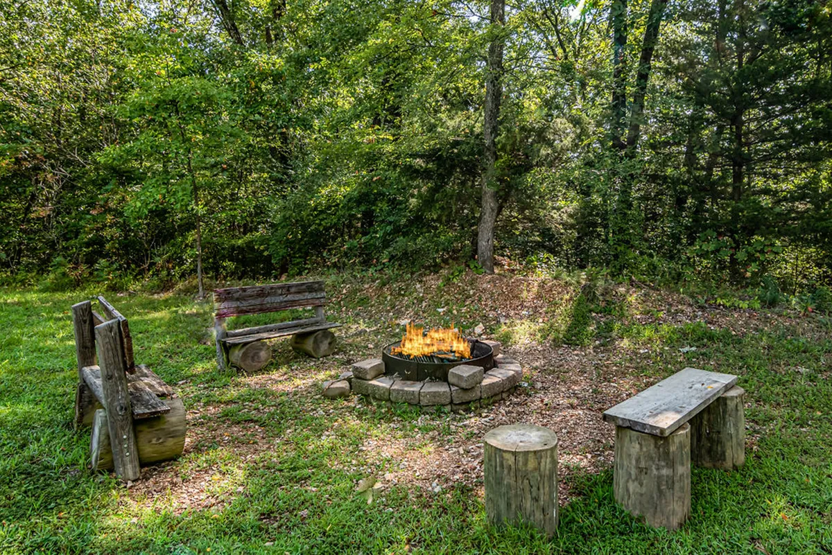 A rustic open-air fire pit surrounded by wooden benches, nestled in lush greenery. The area feels intimate and secluded, ideal for sharing stories, sipping wine, and stargazing in the evening.