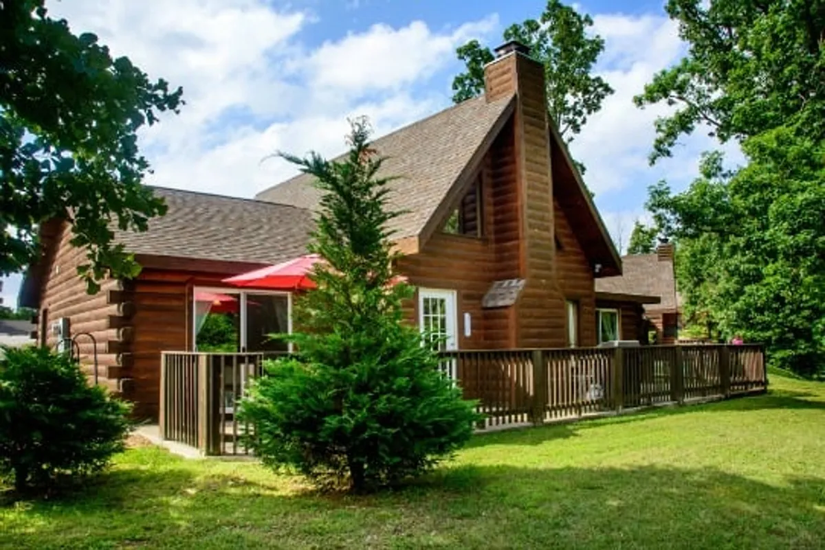 A charming log cabin with a red patio umbrella on a spacious deck, enveloped by green trees. The deck area is set up for outdoor dining and relaxation, embodying the cabin's welcoming, social vibe.