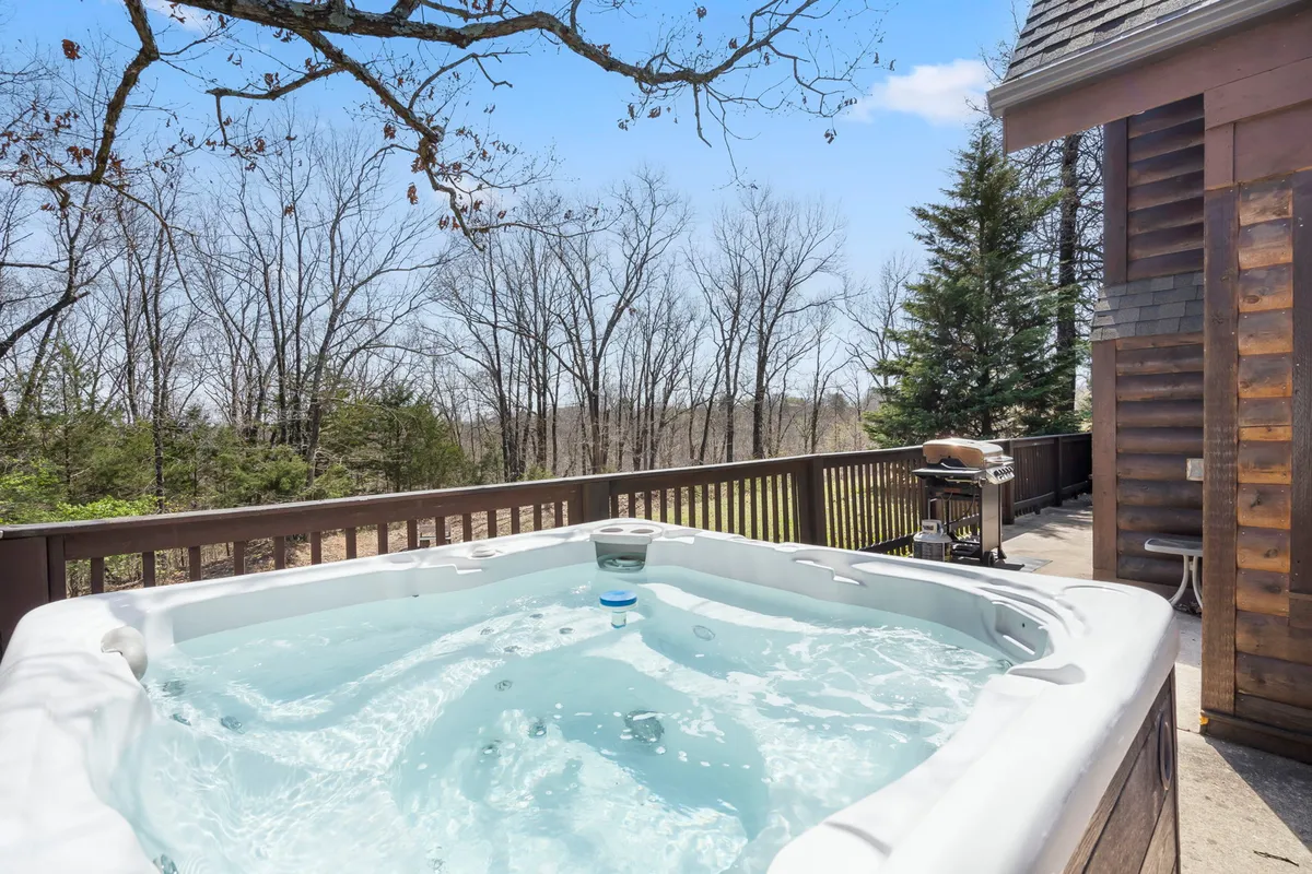 A wooden deck nestled among tall trees, featuring a private hot tub, BBQ grill, and plenty of space for relaxation. The deck is surrounded by lush greenery, offering a tranquil and secluded outdoor setting ideal for unwinding or entertaining.